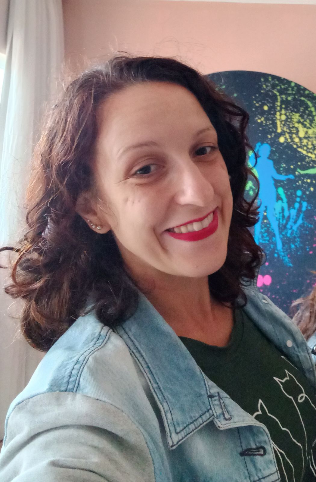 Pessoa adulta com cabelos cacheados castanhos, na altura dos ombros. Está sorrindo, usa batom vermelho, jaqueta jeans clara sobre camiseta escura. A imagem é em ambiente interno, com luz natural e um quadro colorido abstrato ao fundo, transmitindo um clima leve e acolhedor.