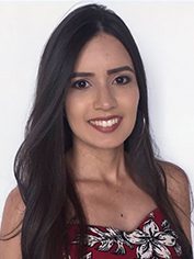 Pessoa com cabelos longos, lisos e escuros, repartidos de lado. Tem expressão sorridente, aparência cuidadosa e usa blusa sem mangas com estampa floral. O fundo é claro e neutro, destacando o rosto e o tronco.