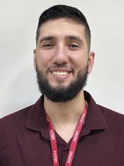 Pessoa com cabelos curtos e escuros e barba cheia bem aparada. Está sorrindo, transmite uma expressão acolhedora e veste camisa polo bordô, usando crachá com cordão vermelho. O fundo é claro e simples.