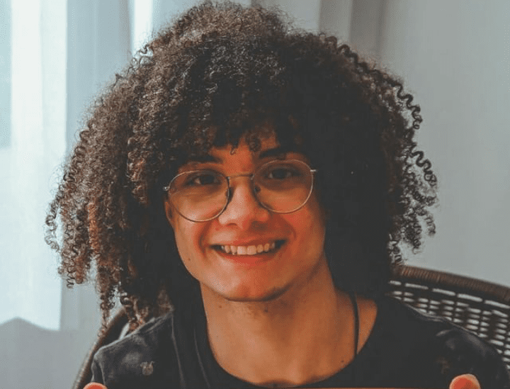 Pessoa jovem, com cabelos cacheados volumosos e escuros, usando óculos de armação redonda. Tem expressão sorridente, aparência amigável e está sentada em um ambiente interno bem iluminado, vestindo camiseta escura.