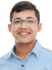 Pessoa jovem com cabelos curtos e escuros, usando óculos de armação fina. Está sorrindo, com expressão aberta e amistosa, e veste camisa social azul-clara. O fundo é claro e neutro.