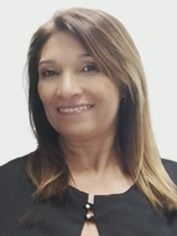 Pessoa adulta com cabelos longos, lisos e castanhos, com mechas mais claras. Tem expressão sorridente, aparência serena e veste blusa preta, com detalhe discreto no decote. O fundo é claro e simples.