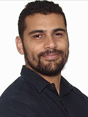 Pessoa adulta com cabelos curtos e escuros e barba cheia bem cuidada. Tem expressão confiante, leve sorriso no rosto e veste camisa escura. O fundo é claro, destacando o rosto e o tronco.